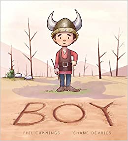 The cover of the book "Boy" features the title and the authors names, Phil Cummings and Shane Devries, below an illustrated smiling boy standing on a barren ground with bare trees behind him. The boy is wearing a Viking helmet, a red long-sleeved shirt with a brown vest and belt, blue pants, and brown shoes.
