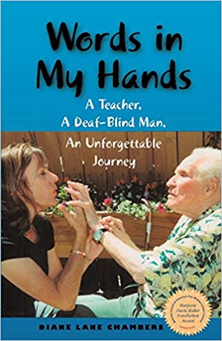 The cover of the book "Words in My Hands: A Teacher, A Deaf-Blind Man, An Unforgettable Journey" features a blue background with the text across the top. Below, an outside setting with a planter box in the background features an old man with his hands on top of a young woman's hands on the left who is signing near her face.