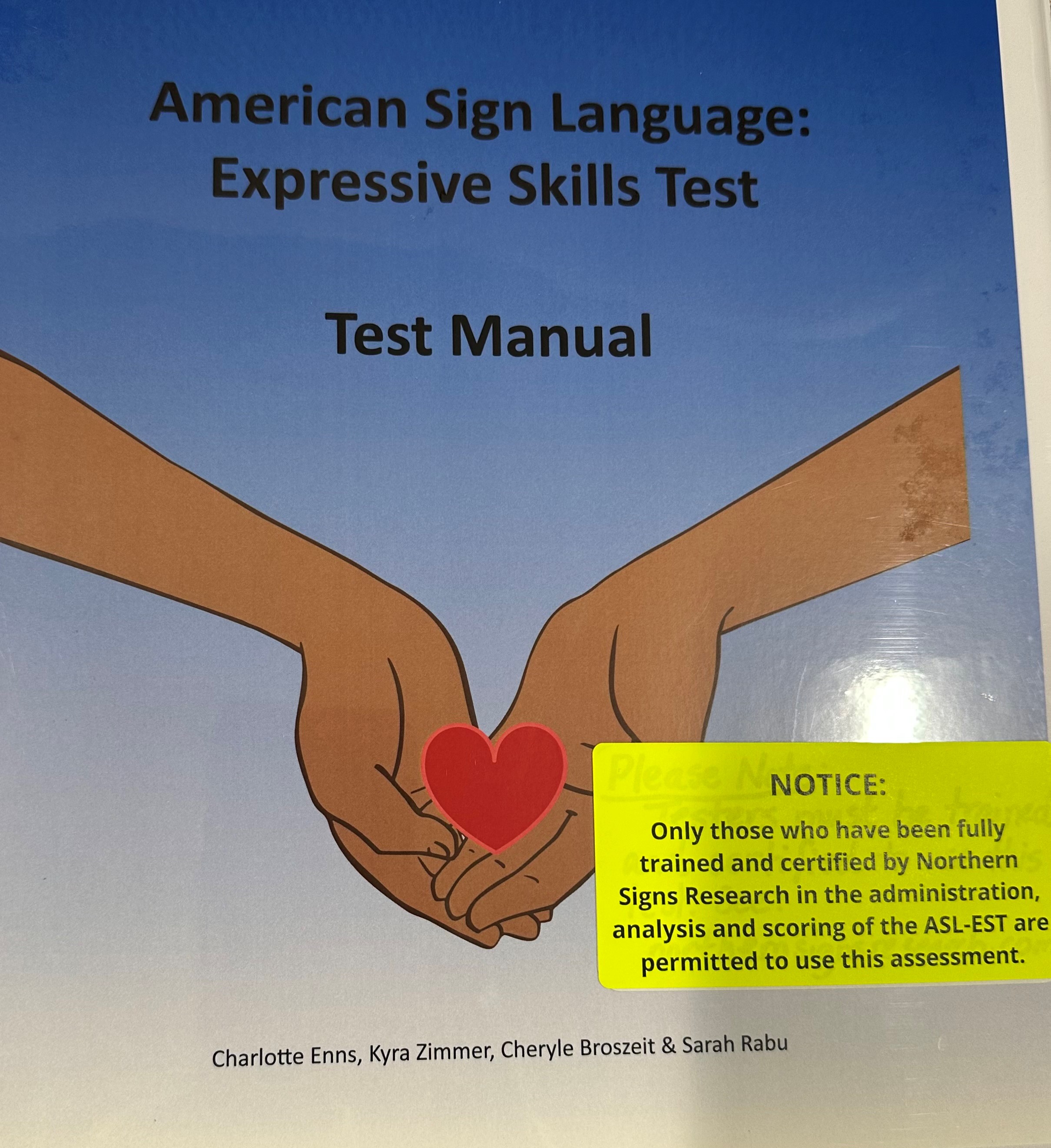 The manual cover for the "American Sign Language: Expressive Skills Test" features a blue background with the title at the top. Below are two illustrated arms coming together and holding a heart in cupped palms.