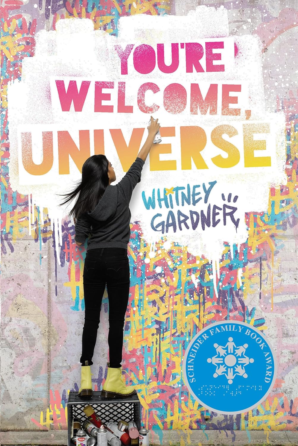 The cover of the book "You're Welcome, Universe" by Whitney Gardner features the image of a girl standing on a crate facing a wall and spray painting the title of the book with additional random graffiti surrounding the wall.