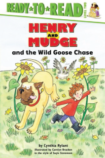 A boy and a dog running past sun flowers. A goose is running behind them.